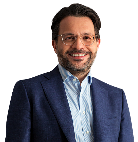 Sócio Gil Mendes está vestindo um paletó azul marinho e camisa azul claro. Ele tem cabelo liso, curto e castanho escuro. Ele está com um largo sorriso.