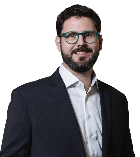 O sócio Luiz Goedert usa óculos redondo de armação verde, tem barba cheia e cabelos pretos, e veste uma camisa branca e um paletó preto.
