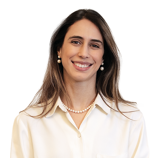 Marcela é uma mulher branca com cabelo loiro escuro. Ela usa uma blusa branca e colar e brincos de pérolas.