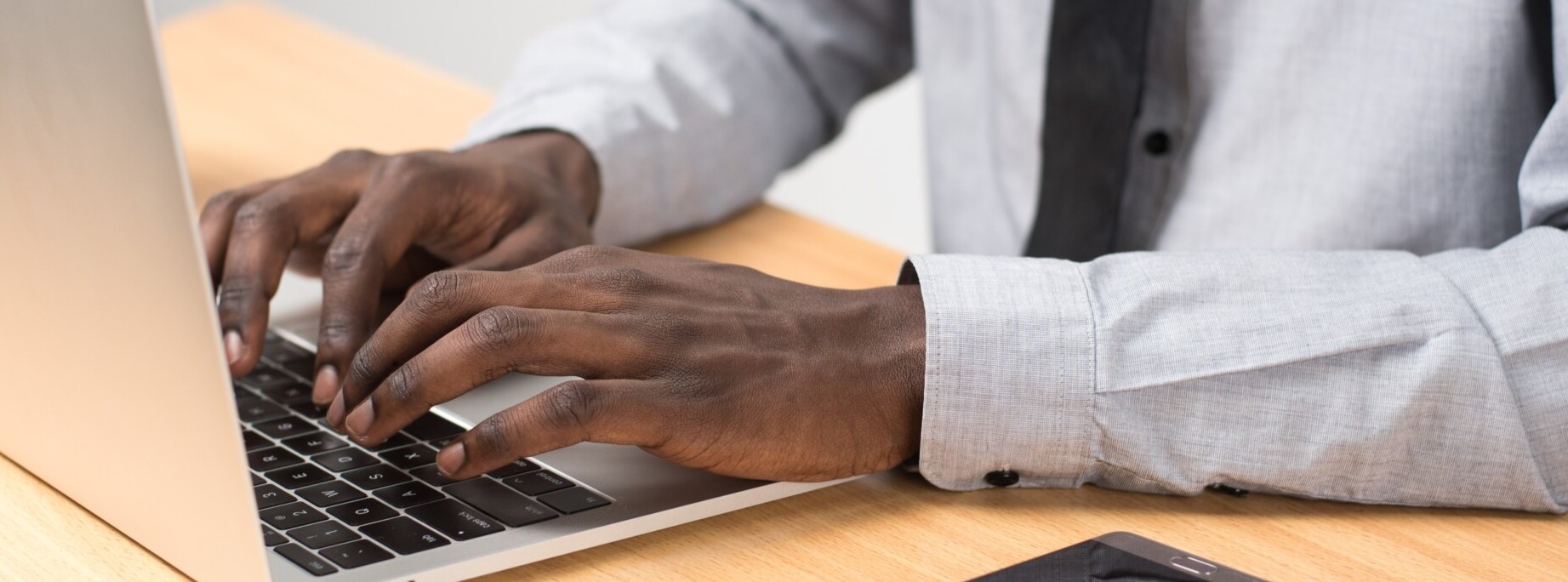 homem de terno e negro manuseia um notebook apoiado em uma mesa de madeira e com um celular ao lado