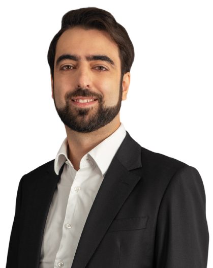 Adriano veste um paletó preto com camisa social branca. Seus cabelos e barba são castanho escuro. Na foto, está com um sorriso.