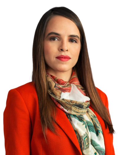 A sócia ariane possui cabelos longos, castanhos, com as pontas mais claras. Na foto, está com uma expressão leve e alegre. Está usando blazer vermelho, com lenço estampado, e batom rosa.