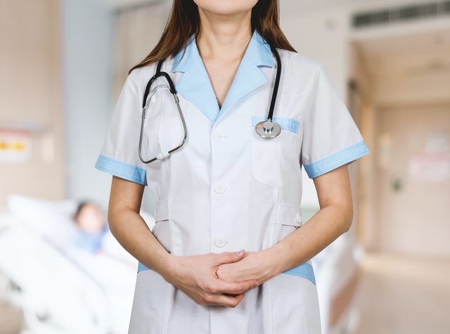 Uma enfermeira de pé usando um uniforme branco e azul e com um estetoscópio no pescoço, e uma cama de hospital com um paciente ao fundo