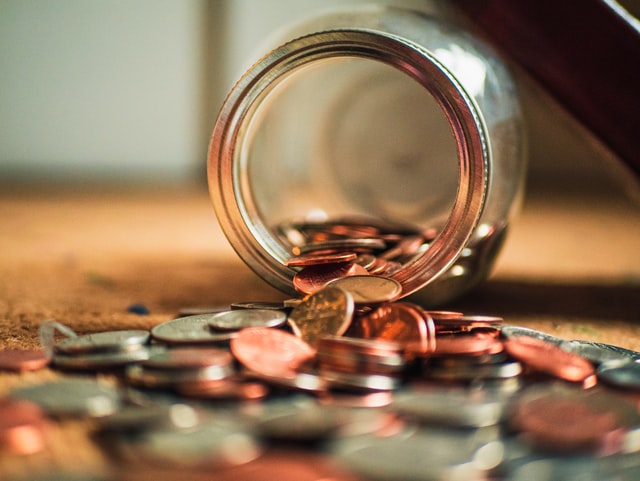 Um pote de vidro caído em uma mesa, com moedas douradas e prateadas saindo do pote