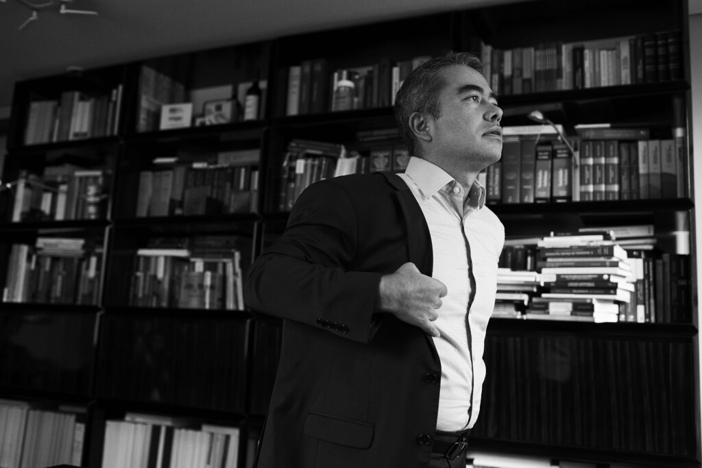 O sócio Jean Arakawa está na biblioteca do escritório de perfil e olhando para cima. Ele está vestindo uma camisa branca e vestindo um paletó escuro. Ele tem cabelo liso e grisalho.