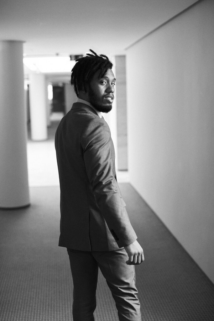 O colaborador Marco Aurelio Farias Duarte está caminhando de costas no corredor do escritório e olhando para trás. Ele veste um terno claro, usa barba e cabelo preto com dreadlock.