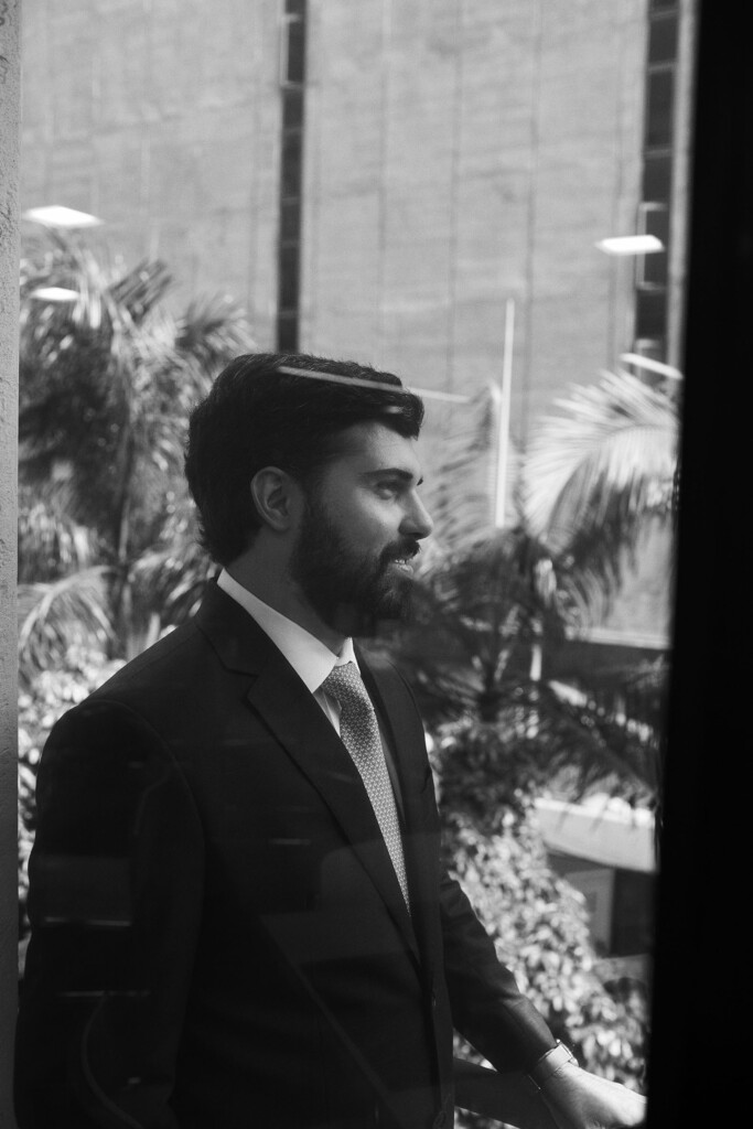 O colaborador Marianno Carneiro está atrás de uma janela de vidro com plantas ao fundo. Ele está de perfil e vestindo um paletó escuro com camisa clara e gravata. Ela usa barba, tem cabelo curto e preto e está com um sorrido com boca fechada.