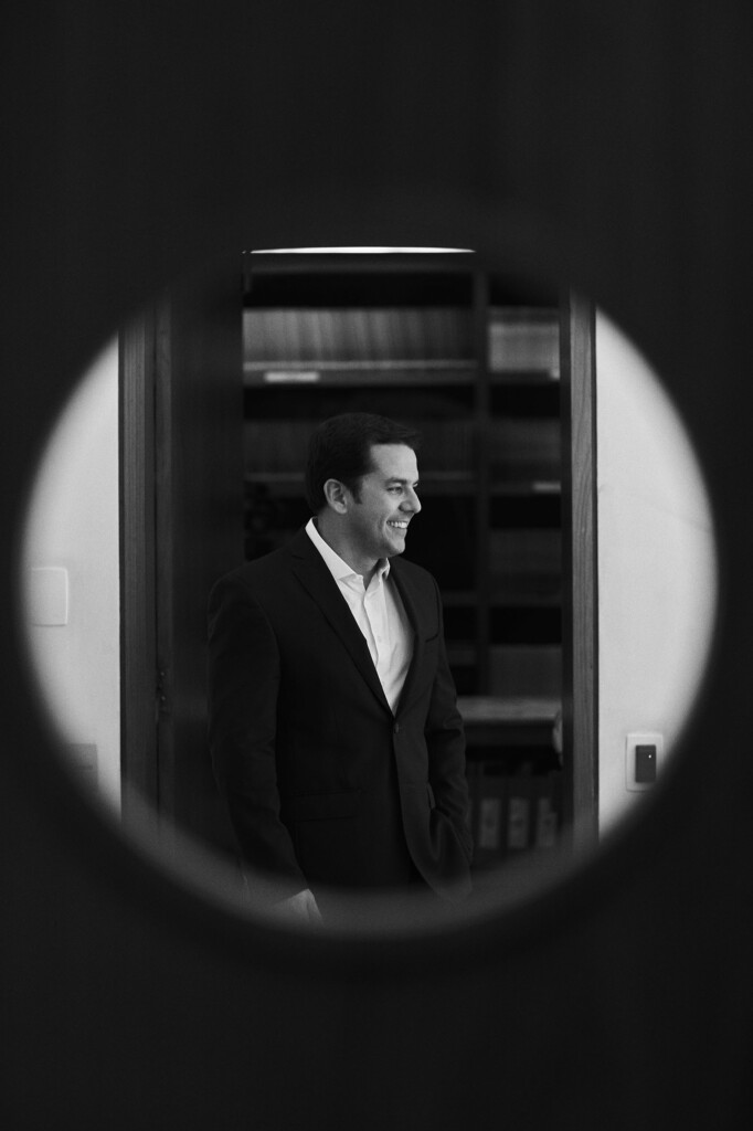 Através de uma clarabóia em uma porta, vimos o sócio Thiago Sombra de perfil em frente à porta da biblioteca. Ele está vestindo um paletó escuro e uma camisa clara. Ele está olhando para o lado e sorrindo.