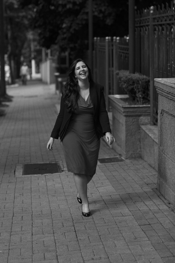 A colaboradora Vivian Falcao está caminhando na calçada do escritório. Ela veste um vestido que vai até abaixo do joelho e um paletó mais escuro com sapatilha. Ela está sorrindo e está com os cabelos soltos, liso e soltos.
