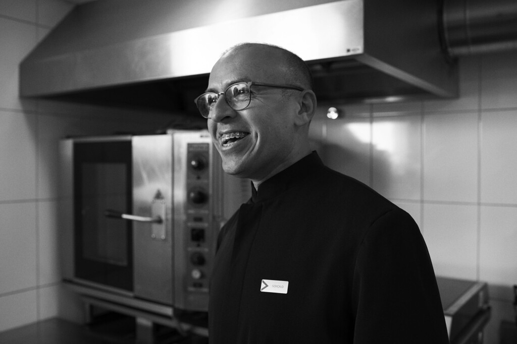 O colaborador Gonçalo Alves da Silva está na cozinha do escritório, em frente à um forno industrial. Ele está vestindo uma camisa preta com um crachá metálico. Ele usa óculo, é careca e está sorrindo.
