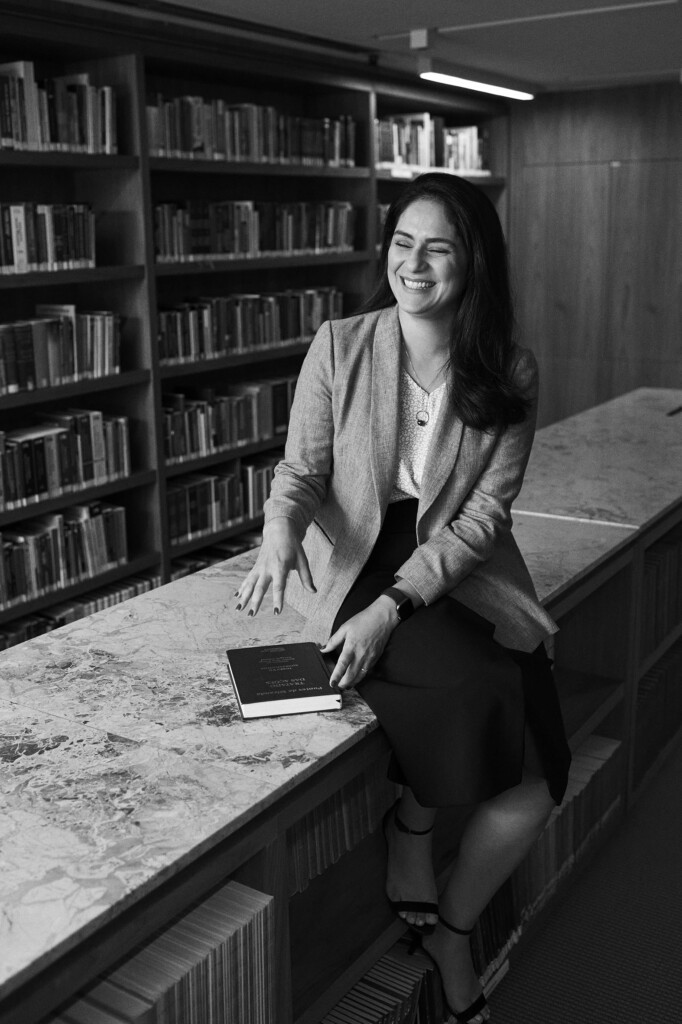 Na biblioteca do escritório, a advogada Joana Pimentel Gomes, está sentada em uma bancada, sorrindo e com um livro em suas mãos. Ela veste uma roupa social e tem cabelos longos e escuro. A foto está em preto e branco.