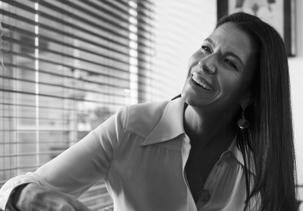 No escritório, a sócia Marina Anselmo, está super sorridente em sua sala. Ela veste uma camisa social clara de manga longa. Ela tem cabelos longos e escuros. A foto está em preto e branco.