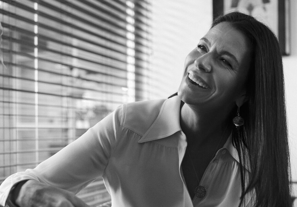 No escritório, a sócia Marina Anselmo, está super sorridente em sua sala. Ela veste uma camisa social clara de manga longa. Ela tem cabelos longos e escuros. A foto está em preto e branco.