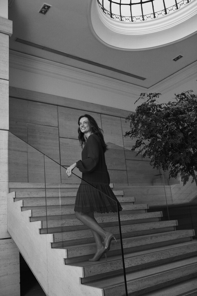 No escritório, a sócia Vanessa Fiusa, está subindo umas escadas de mármore. Ela está usando um vestido longo e escuro. Ela tem cabelos longos, ondulados e com mechas. A foto está em preto e branco.