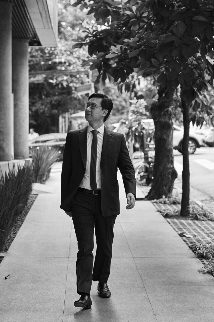 O sócio alex hatanaka está caminhando pela calçada nos arredores do escritório. Ele está vestindo paletó, calça social e gravata em tom escuro, e blusa social em tom claro. A foto está em preto e branco.