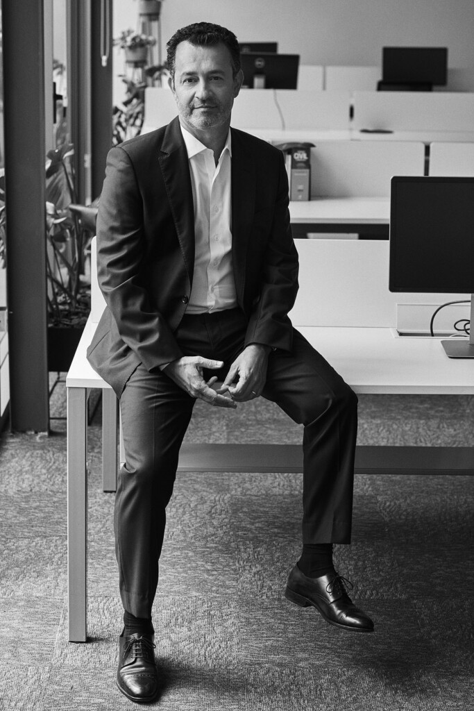 O sócio eduardo damião está no escritório, sentado sobre a mesa, ao lado do computador. Ele está vestido paletó e calça em tom escuro, e blusa social em tom claro. A foto está em preto e branco.