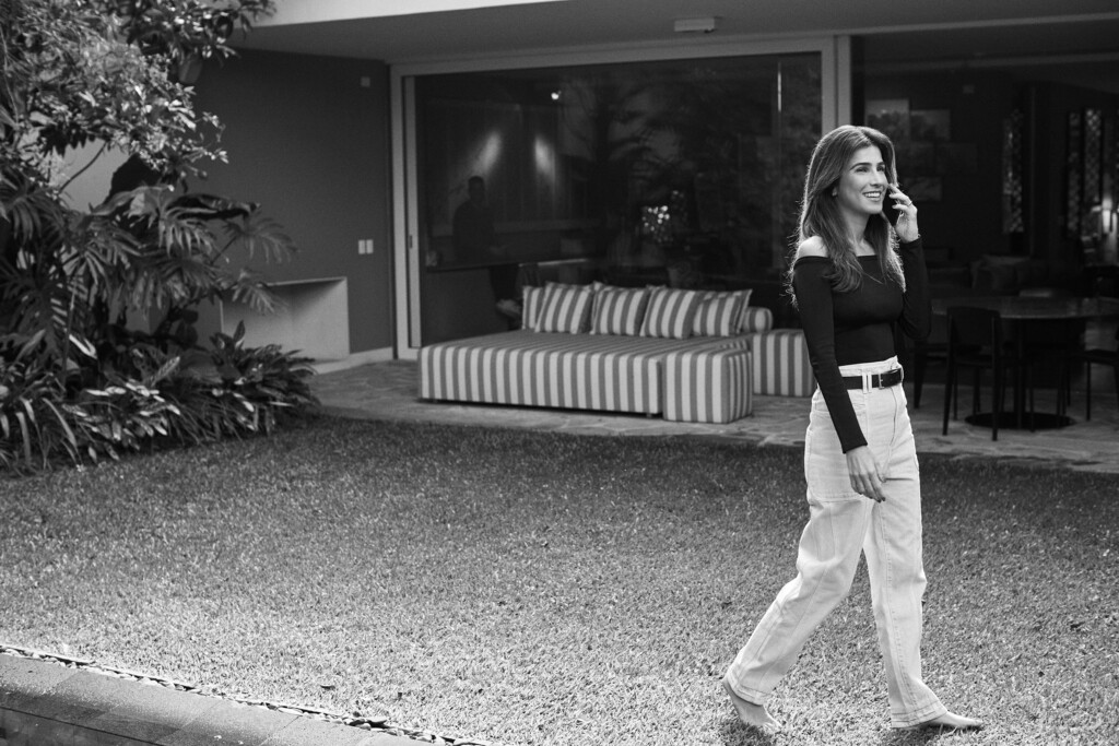 A sócia Marina Procknor está em um momento de lazer, andando em sua casa. Ela é morena, tem cabelos longos e veste uma calça clara e blusa de maga longa escura. A foto está em preto e branco.
