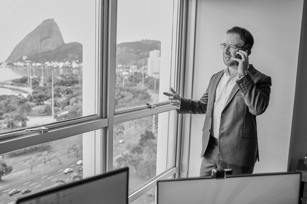 O sócio Claudio Oksenberg está em sua sala, em pé e falando ao telefone. Ele está perto da grande janela de sua sala, onde é possível ver o Pão de Açúcar, e ele aprecia a vista. Ele está vestido com um Blazer e uma calça de alfaitaria, e uma blusa de botão branca e de manga longa. Ele é branco, usa óculos, tem o cabelo castanho claro curto e a barba cerrada. Ele gesticula com as mãos. É possível ver uma parte das telas de seu computador na sala. A foto é preta e branca.