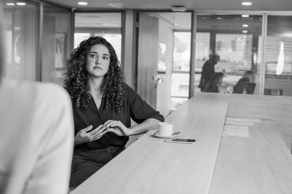 A advogada Fernanda Maranhão está sentada e sobre a mesa, há um café e um celular. Ela olha para o lado e a ponta dos seus dedos se encontram. Ela está vestida com uma blusa de manga longa e de botão e uma calça. Ela é branca e tem o cabelo castanho, longo e cacheado. A foto é preta e branca.