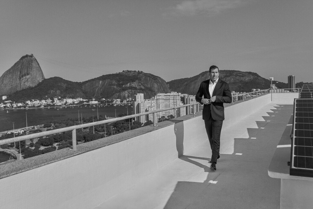 No último andar do escritório do Rio de Janeiro, com montanhas ao fundo, o sócio Pablo Sorj está caminhando, de blazer e calça social. Ele é alto, com estrutura física larga e tem o cabelo curto e é branco. Ele está olhando para o lado e está abotoando o blazer. A foto é preta e branca.