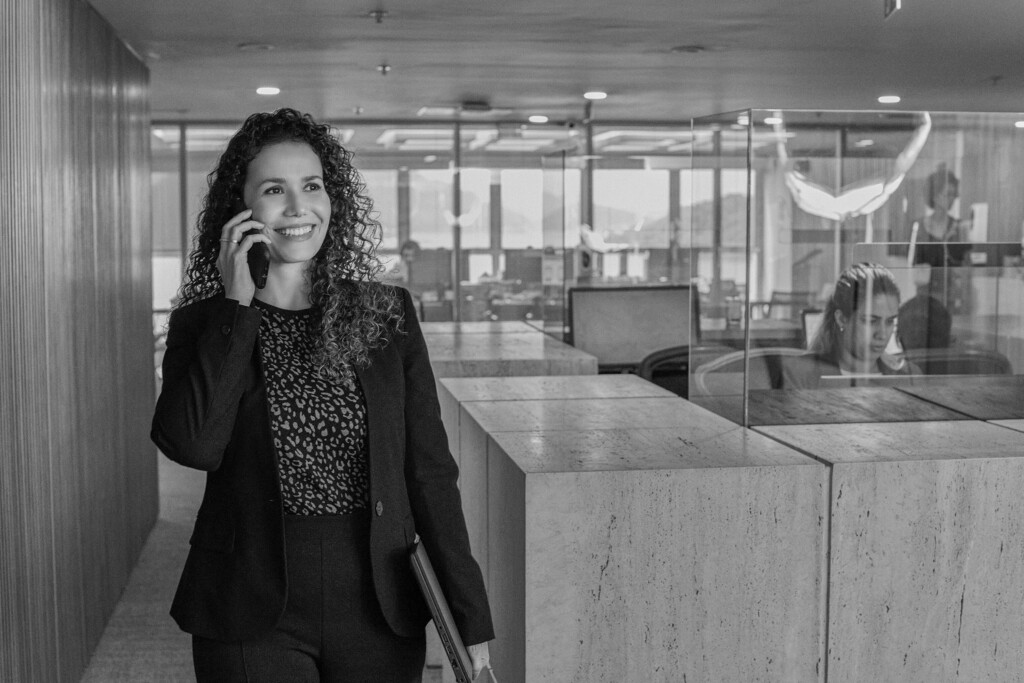 A colaboradora Priscilla de Souza está com um largo sorriso no rosto, fala ao telefone e segura um notebook com a mão esquerda. Ela está na recepção do andar onde contém as salas de reunião. Ela veste um blazer e calsa de alfaitaria preta e uma blusa estampada de oncinha. Ela é branca e tem o cabelo longo e cacheado e castanho. A foto é preta e branca.