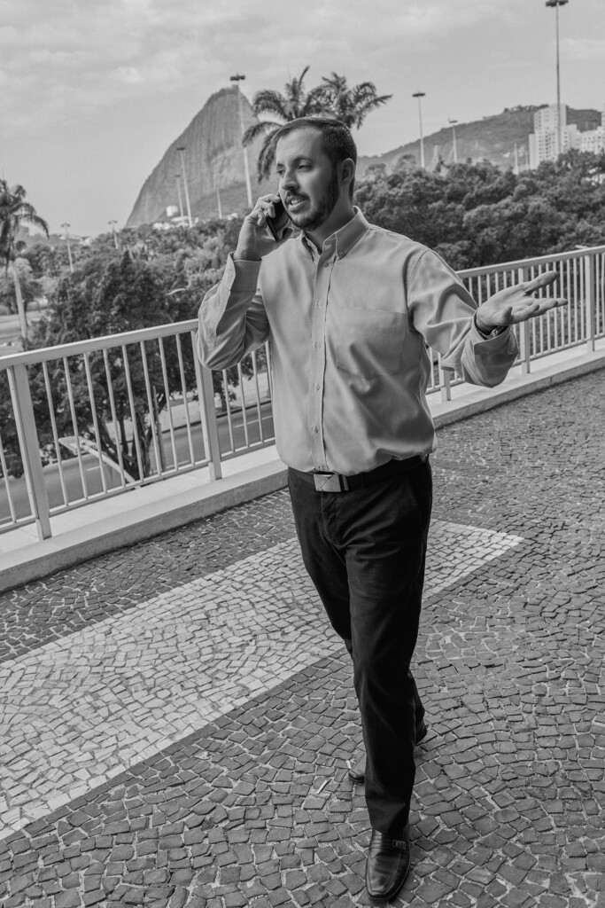 O colaborador sergio nagel está caminhando e falando ao telefone na área externa do escritório, com vista para o pão de açúcar, no rio de janeiro. Ele está vestido calça escura e camisa social clara. a foto está em preto e branco.
