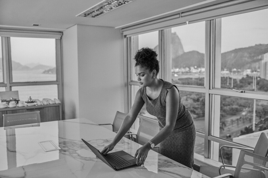 A colaboradora yasmin carneiro está em uma sala de reunião com amplas janelas e vista para o pão de açúcar, no rio de janeiro. Usando um vestido, ela está colocando o notebook em cima da mesa de mármore. A foto está em preto e branco.