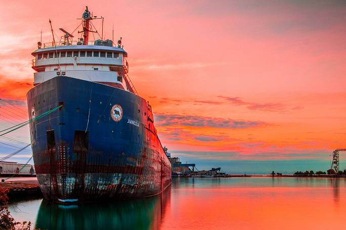 Navio cargueiro azul escuro atracado. Na imagem, destaca-se o pôr do sol que deixa as nuvens no céu em tons de rosa e laranja, fazendo um reflexo da mesma cor na água do mar,
