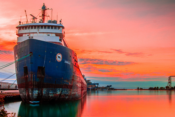 Dark blue cargo ship docked. In the image, the sunset stands out, leaving the clouds in the sky in shades of pink and orange, making a reflection of the same color in the sea water,