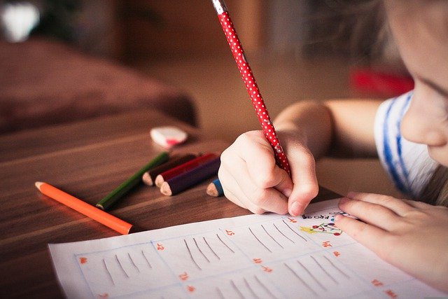 Uma criança escrevendo em um papel com um lápis vermelho, também há uma borracha e lápis de várias cores em cima da mesa