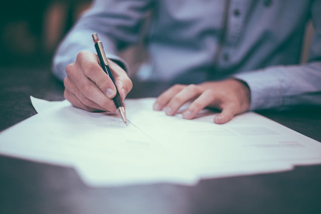 A man in a blue shirt writing on a paper on top of a brown table