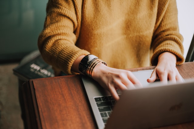 pessoa de suéter amarelo mostarda digitando em um notebook que está sobre uma mesa marrom