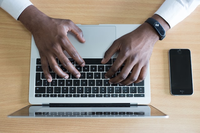 Mãos de um homem negro digitando em um notebook, há um celular ao lado