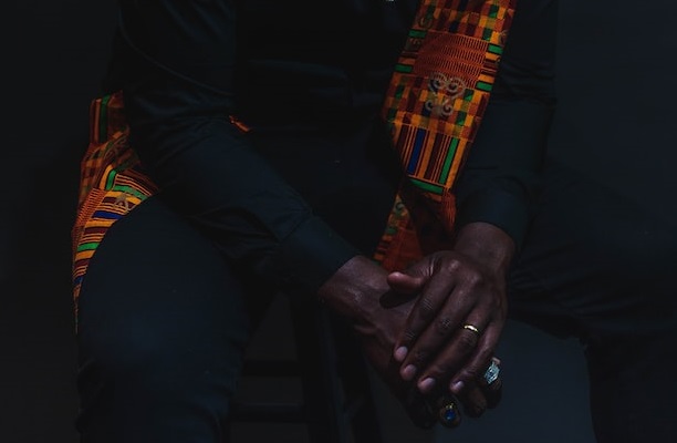 imagem com foco no tronco e nas mãos de um homem negro sentado, com destaque para uma faixa africana colorida contrastando com o resto da imagem