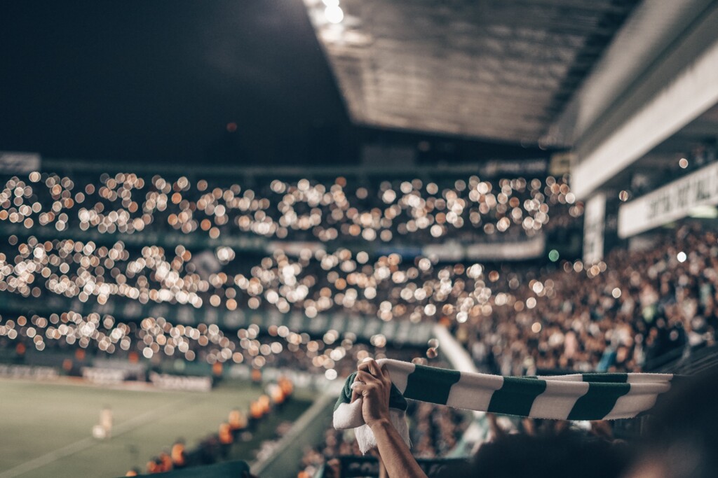 Estádio de futebol, com a torcida com as luzes dos celulares acesas e uma pessoa segurando um cachecol verde e branco nas mãos