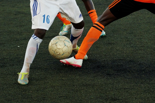 Dois homens negros jogando futebol. Um está com uniforme branco com detalhes em azul e o outro com laranja e detalhe preto.