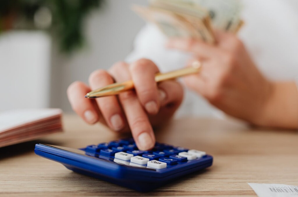mãos de uma mulher branca, sendo que uma está segunda cédulas de dinheiro e a outra está digitando em uma calculadora azul enquanto segura uma caneta