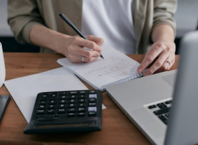 Pessoa calculando em um caderno com um notebook e uma calculadora ao lado