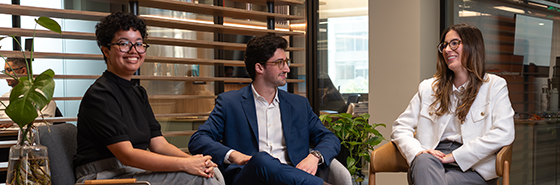 Três pessoas sentadas conversando em cadeiras modernas em um ambiente corporativo. Ao fundo, há divisórias de madeira e uma parede de vidro que revela um espaço de escritório iluminado.