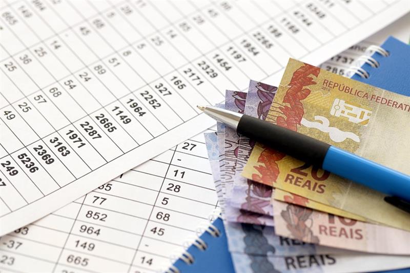 Caneta com reais brasileiros sobre documentos com tabela de cálculos.