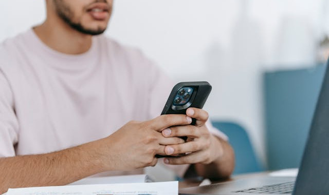 Homem branco mexendo no celular em frente a um notebook